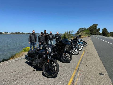 Riders gathered behind several motorcycles beside the water.