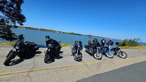 Motorcycles lined up beside the water with riders gathered behind them.