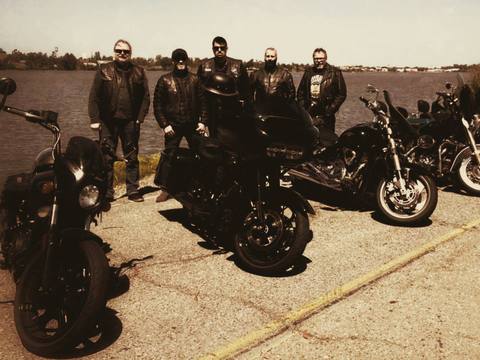 Riders standing with motorcycles beside the water in a warm-toned photo.