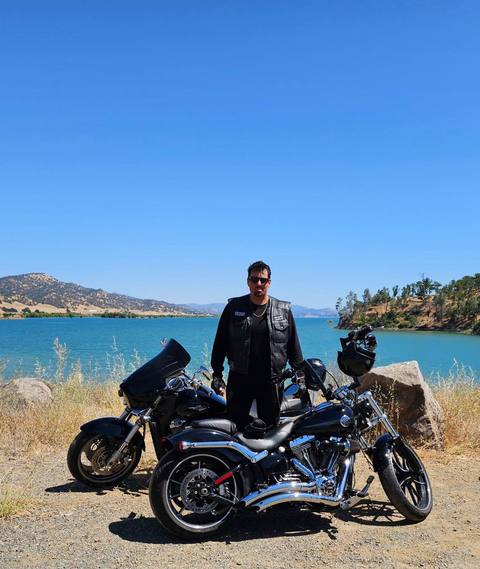 A rider standing beside motorcycles with water and hills behind.