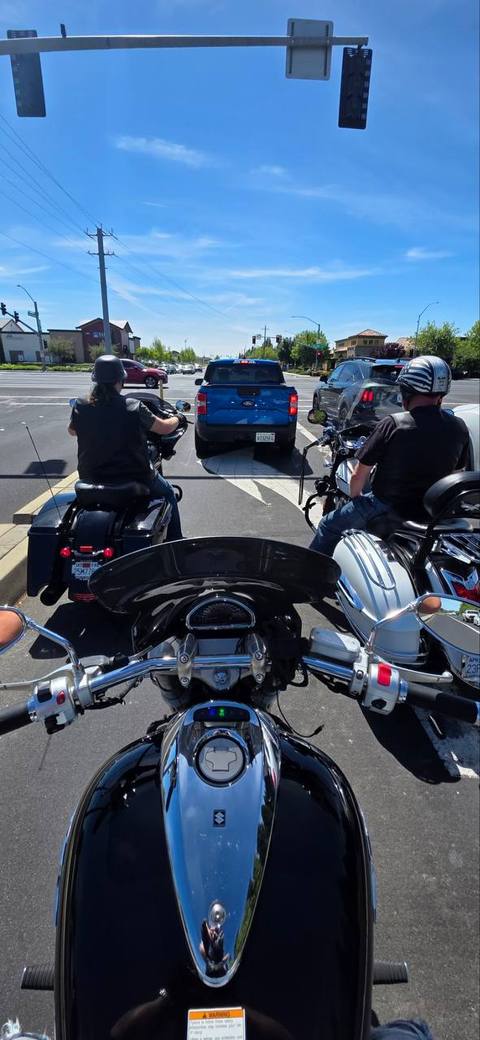 A view over a motorcycle dashboard toward riders waiting in traffic.