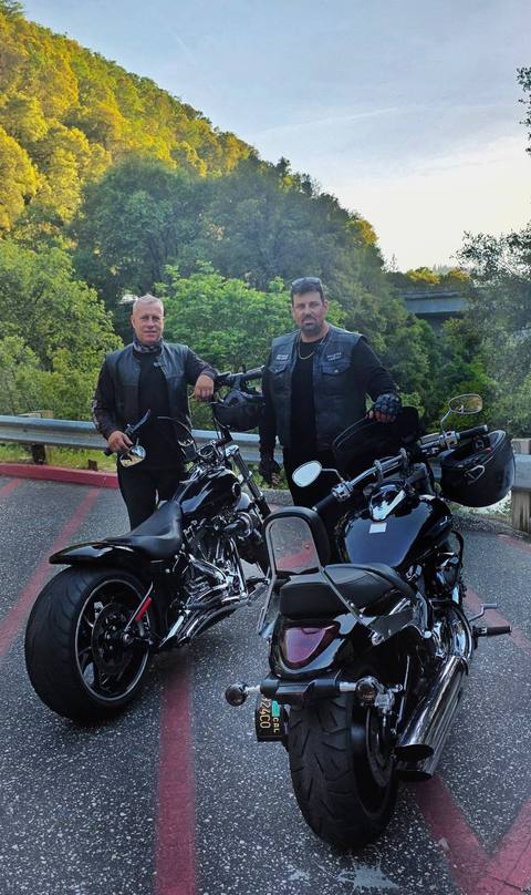 Two riders standing with motorcycles beside a wooded hillside.