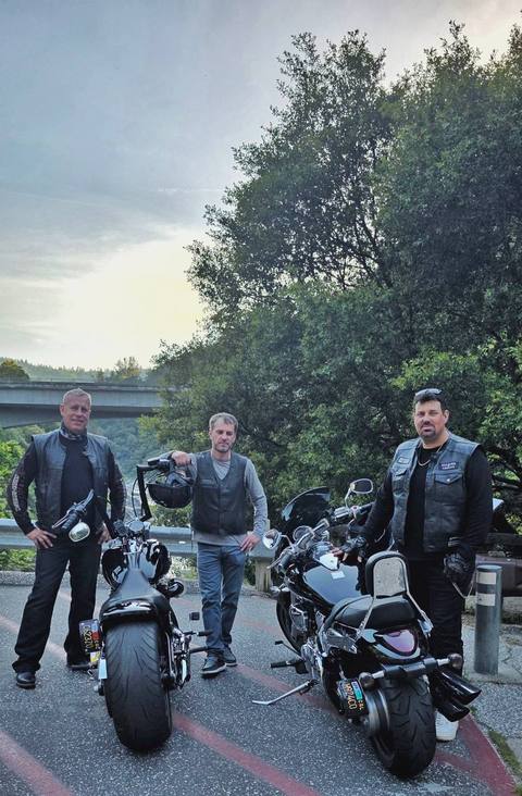 Three riders standing with motorcycles beside a wooded hillside.