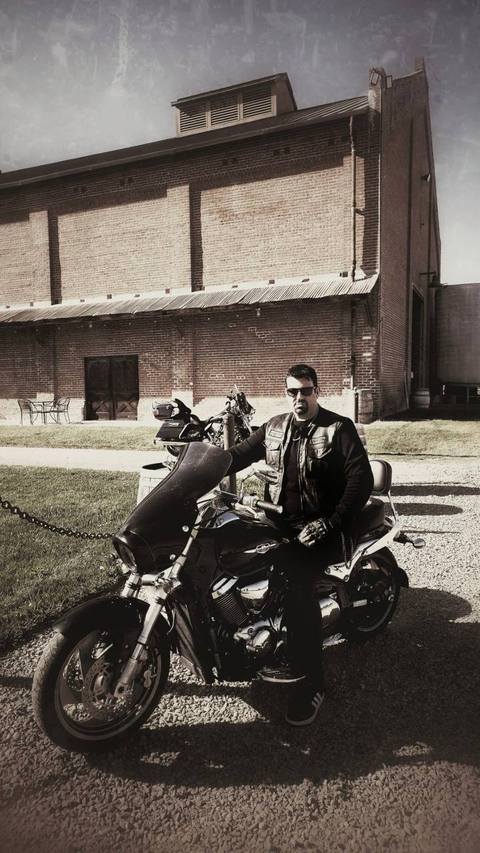 A rider seated on a motorcycle beside a building in a sepia-toned photo.