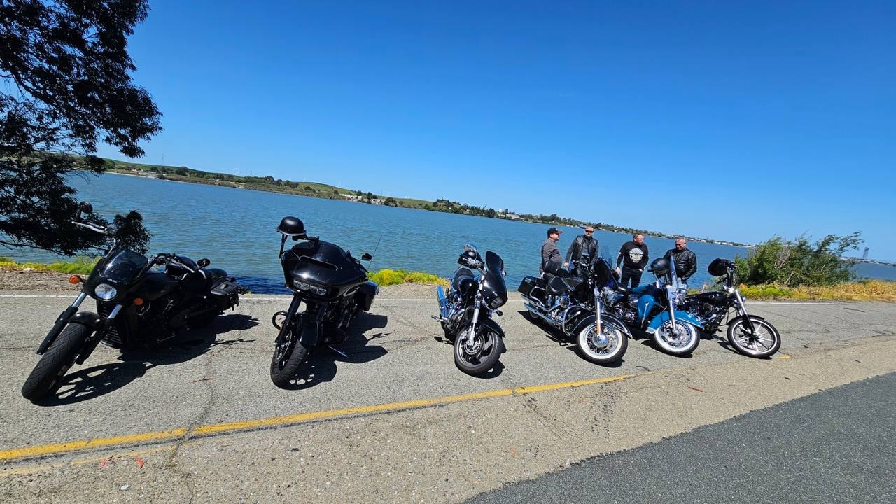 Motorcycles lined up beside the water with riders gathered behind them.