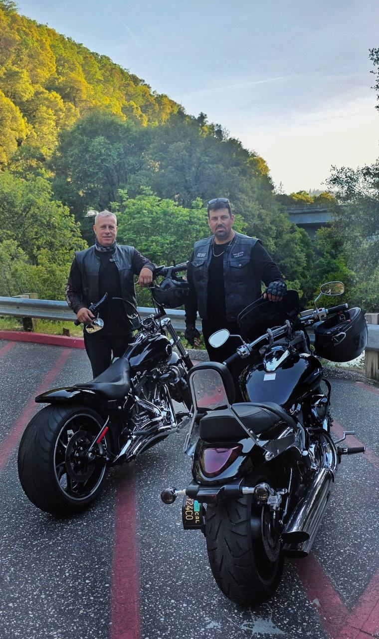 Two riders standing with motorcycles beside a wooded hillside.